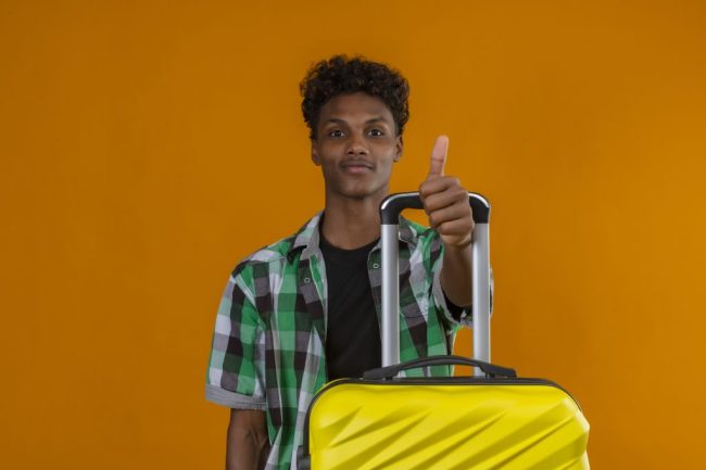 young african american traveler man standing with suitcase looking at camera smiling showing thumbs up over orange background